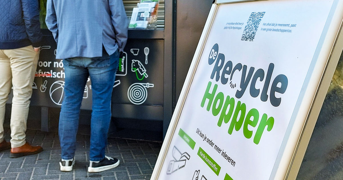 Stoepbord met informatie over De RecycleHopper met op de achtergrond benen van mensen die bij De RecycleHopper staan.